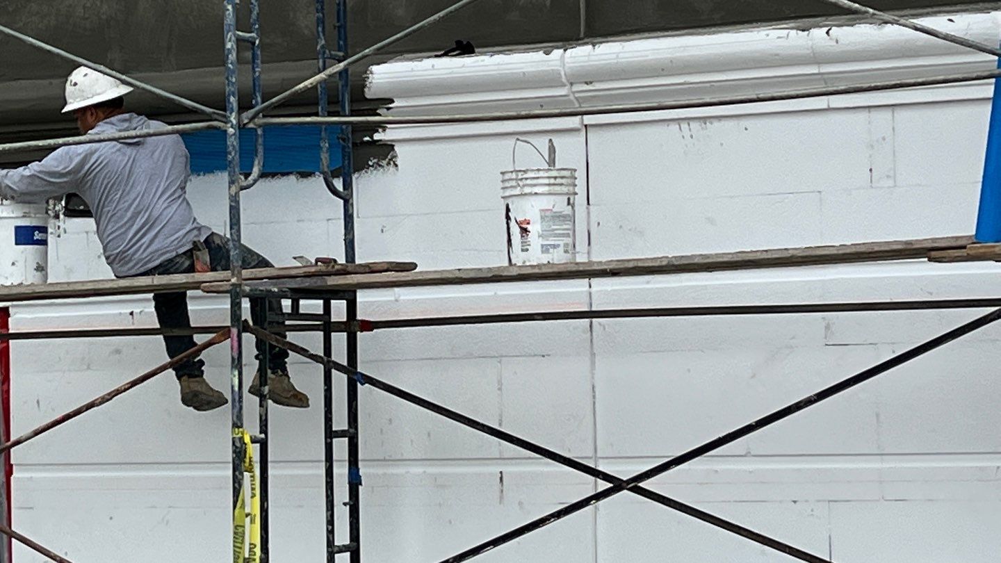 MileHigh worker on scaffolding applying white paint or plaster to a building exterior wall with decorative molding, using buckets of material nearby in westiminster, co
